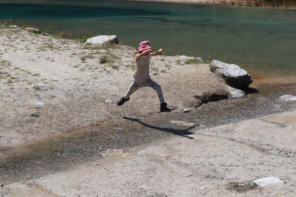 A child wearing a pink hat and light clothing is captured mid-air, jumping playfully on a sandy shore next to clear blue-green water